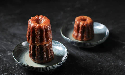 Deux canelés caramélisés disposés dans des coupelles en céramique, l'un empilé sur l'autre au premier plan, avec un second canelé dans une coupelle en arrière-plan, sur une surface noire.