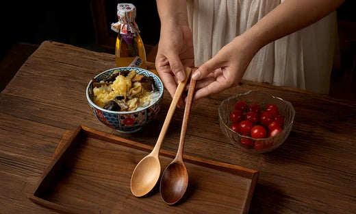 Deux cuillères en bois tenues au-dessus d'un plateau en bois, avec un bol de riz garni de légumes et une salade de tomates cerises sur une table rustique en bois.