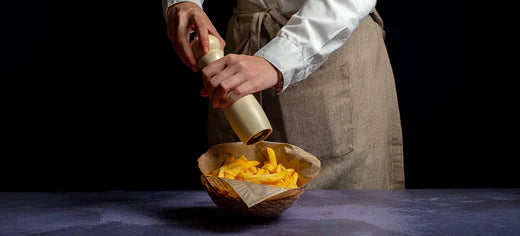 Personne ajoutant du poivre sur des frites croustillantes dans un bol avec un moulin à poivre, portant un tablier beige et une chemise blanche