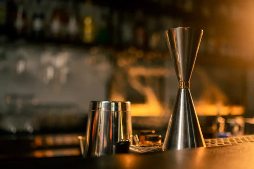  Un comptoir de bar éclairé en lumière chaude avec des outils de mixologie, dont un doseur cocktail métallique et un shaker en acier inoxydable, posés au premier plan.