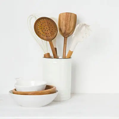 Une collection d'ustensiles de cuisine en bois et en plastique, comprenant une spatule, une cuillère perforée et un fouet, placée dans un pot blanc. Des bols et assiettes en céramique blanche et bois sont à côté.