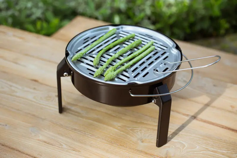 Mini barbecue portable au charbon posé sur une table en bois, grillant des asperges vertes en plein air.