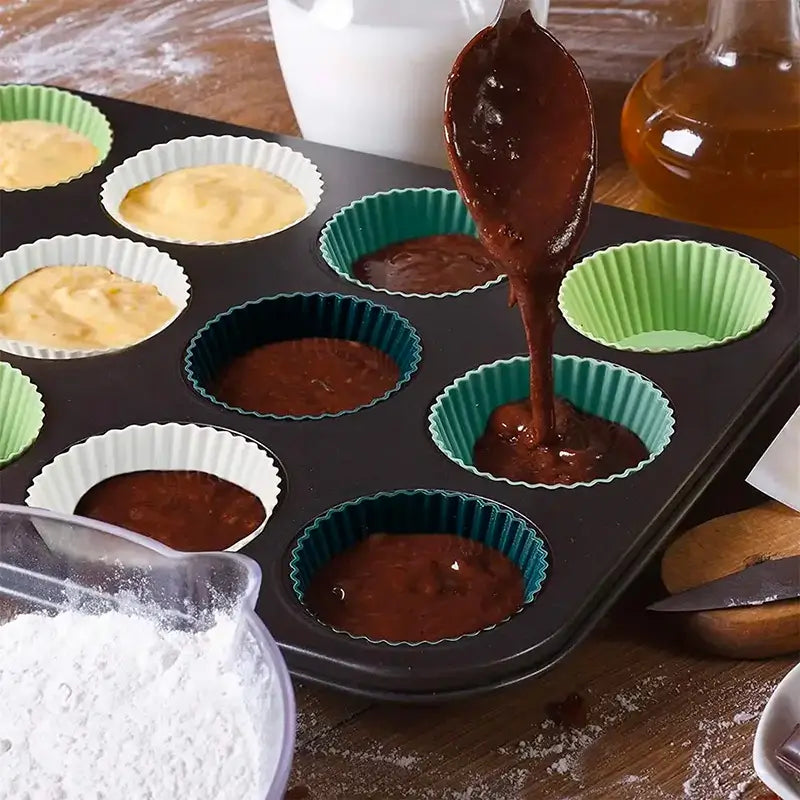 Moules à cupcakes en silicone placés dans un moule métallique, remplis de pâte à cupcakes de différentes couleurs, avec une cuillère versant de la pâte au chocolat.