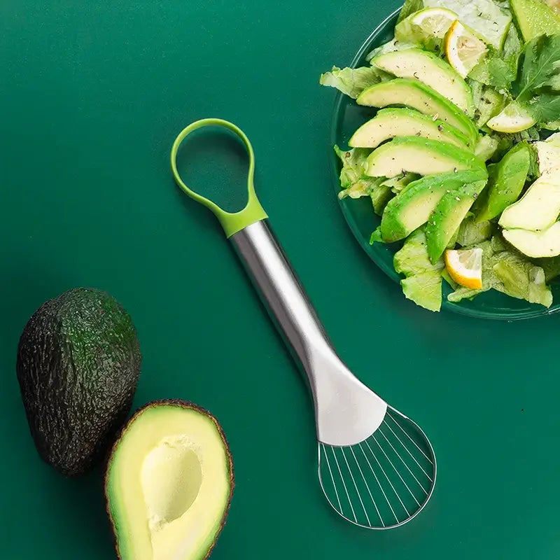 Un ustensile de cuisine en acier inoxydable avec une poignée verte, présenté sur un fond vert avec un avocat et une assiette de salade contenant des tranches d'avocat.