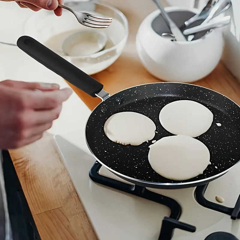 Une poêle contenant trois crêpes en cours de cuisson sur une cuisinière, avec des ustensiles et un bol de pâte en arrière-plan.