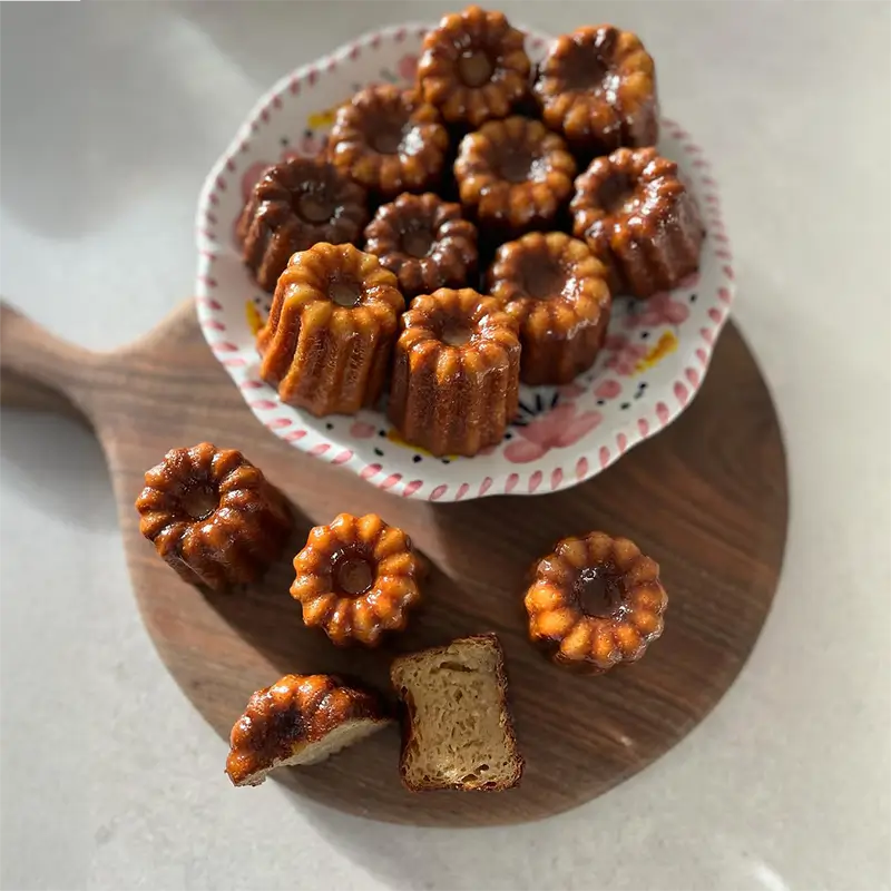 Assiette colorée remplie de cannelés maison dorés et caramélisés, placée sur une planche en bois, avec quelques cannelés posés autour.