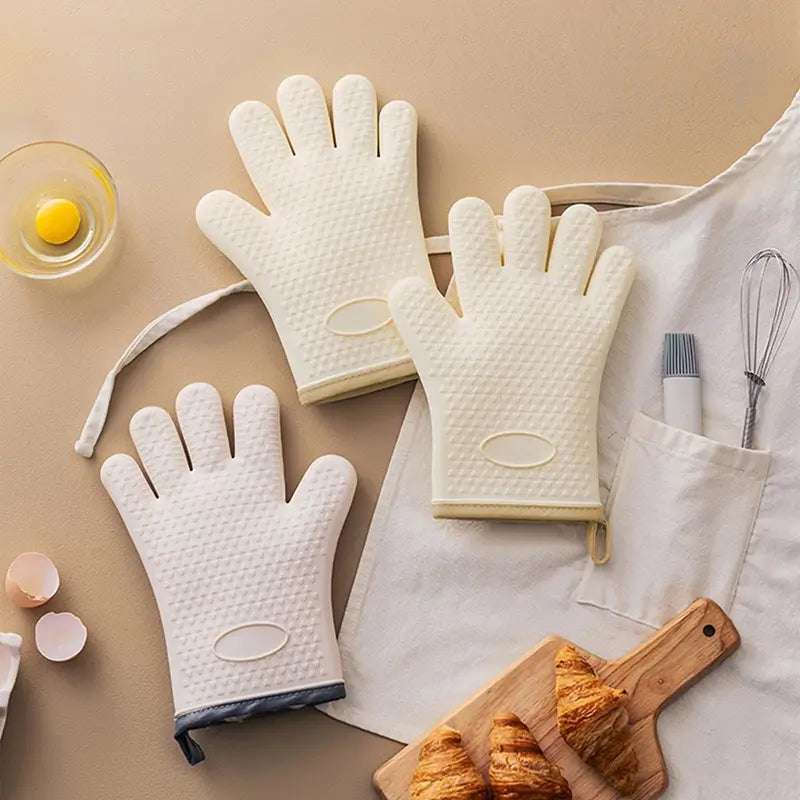 Trois gants de cuisine en silicone blanc et beige disposés sur un plan de travail beige, accompagnés d'accessoires de cuisine tels qu'un fouet, un pinceau, et des croissants sur une planche en bois.