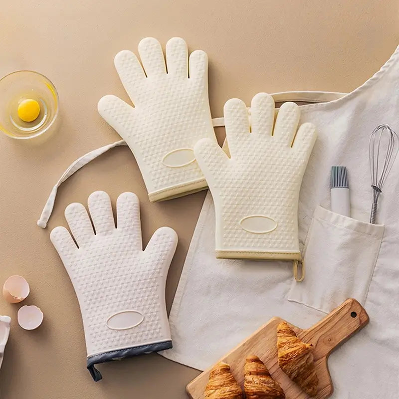 Trois gants de cuisine en silicone blanc et beige disposés sur un plan de travail beige, accompagnés d'accessoires de cuisine tels qu'un fouet, un pinceau, et des croissants sur une planche en bois.