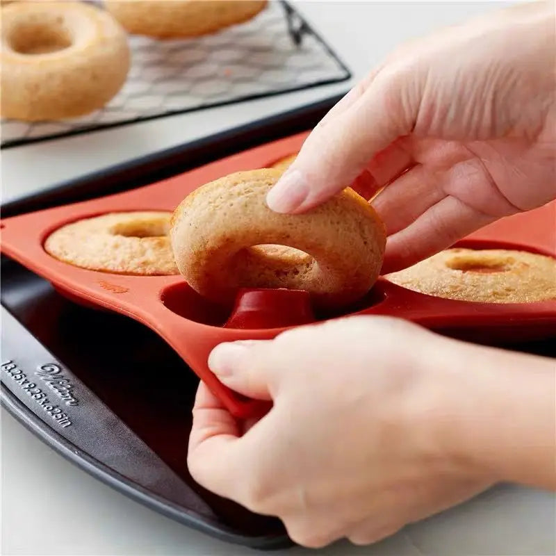 Une main retirant un donut cuit d'un moule en silicone rouge posé sur une plaque de cuisson noire, avec des donuts supplémentaires sur une grille de refroidissement à l'arrière.