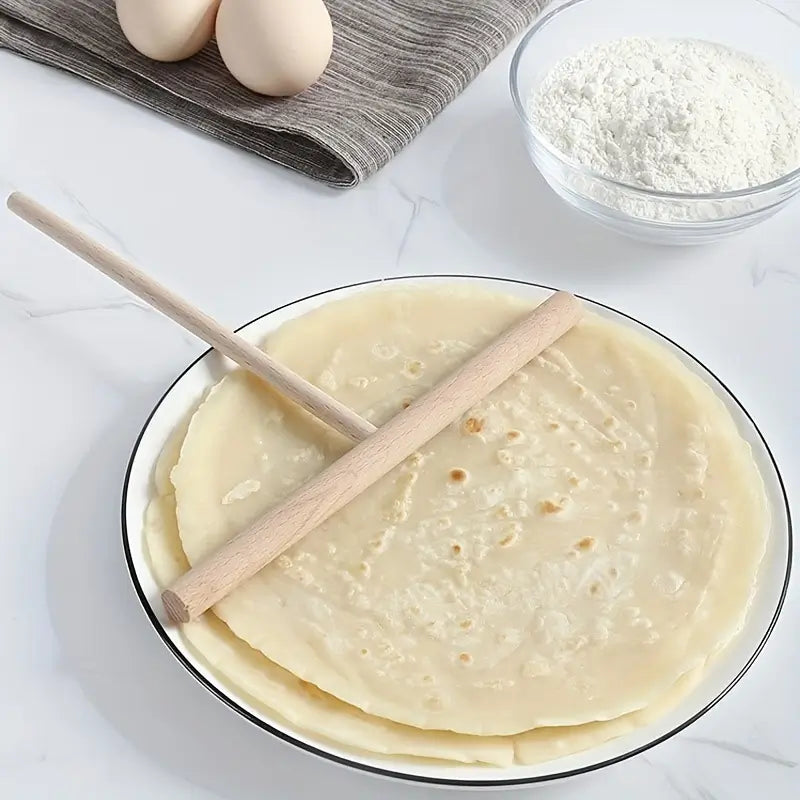 Râteau à crêpes en bois posé sur une pile de crêpes sur une assiette blanche, avec des ingrédients comme des œufs et de la farine en arrière-plan.